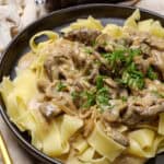 Creamy mushroom and beef stroganoff served over wide egg noodles garnished with chopped fresh chives on a black plate. Rustic background with mushrooms and peppercorns for a hearty, comforting meal.