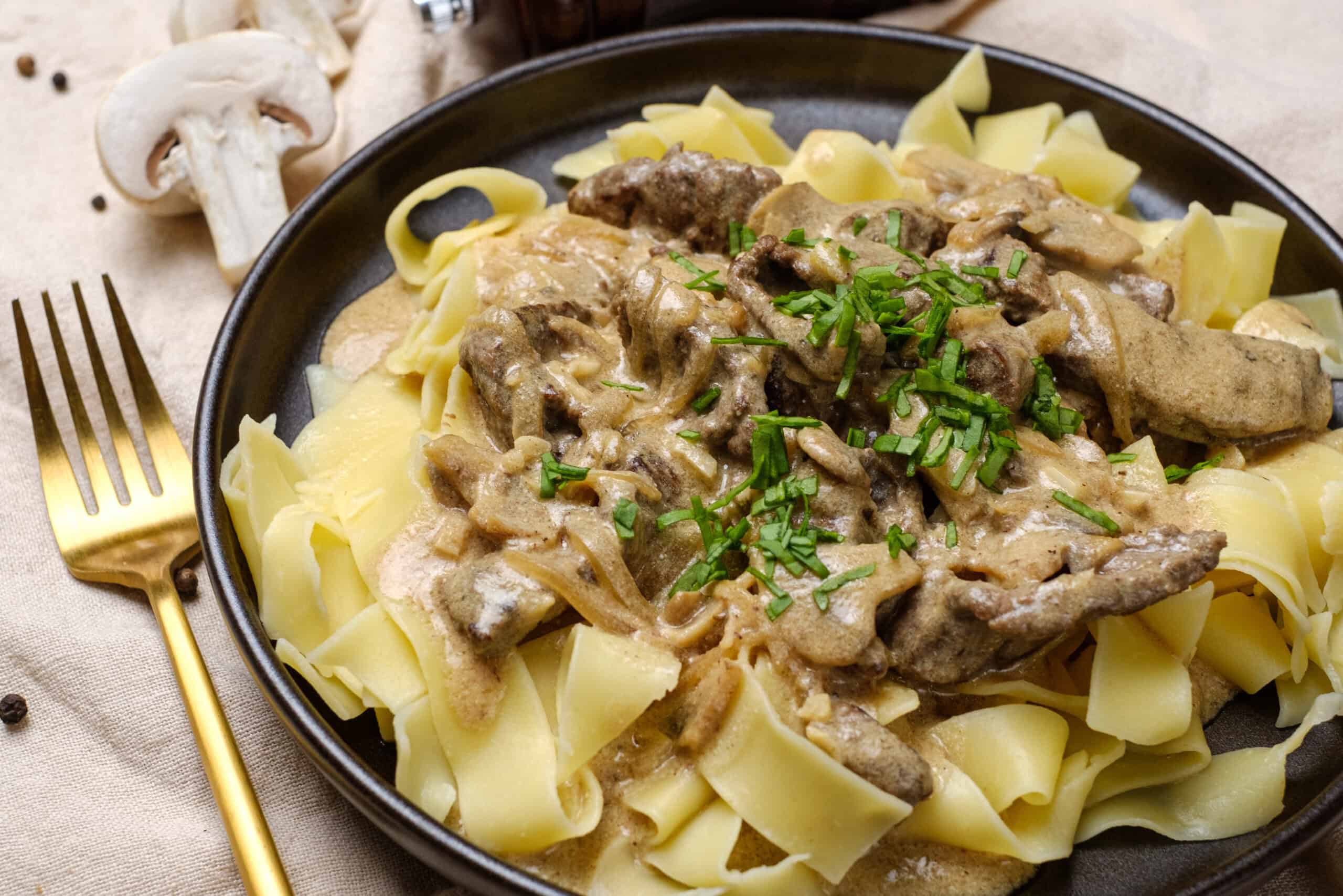 Creamy mushroom and beef stroganoff served over wide egg noodles garnished with chopped fresh chives on a black plate. Rustic background with mushrooms and peppercorns for a hearty, comforting meal.