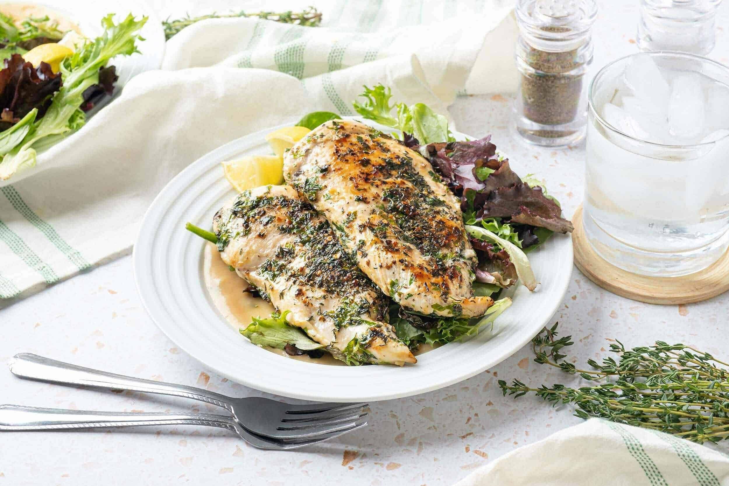 Grilled herb-marinated chicken breasts served on a bed of mixed greens with lemon wedges, accompanied by a glass of ice water, salt, pepper, and fresh thyme for a healthy and flavorful meal.