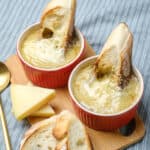 Creamy French onion soup served in red ramekins with toasted baguette slices and melted cheese, garnished with sliced cheese on a wooden board, perfect for cozy comfort food.