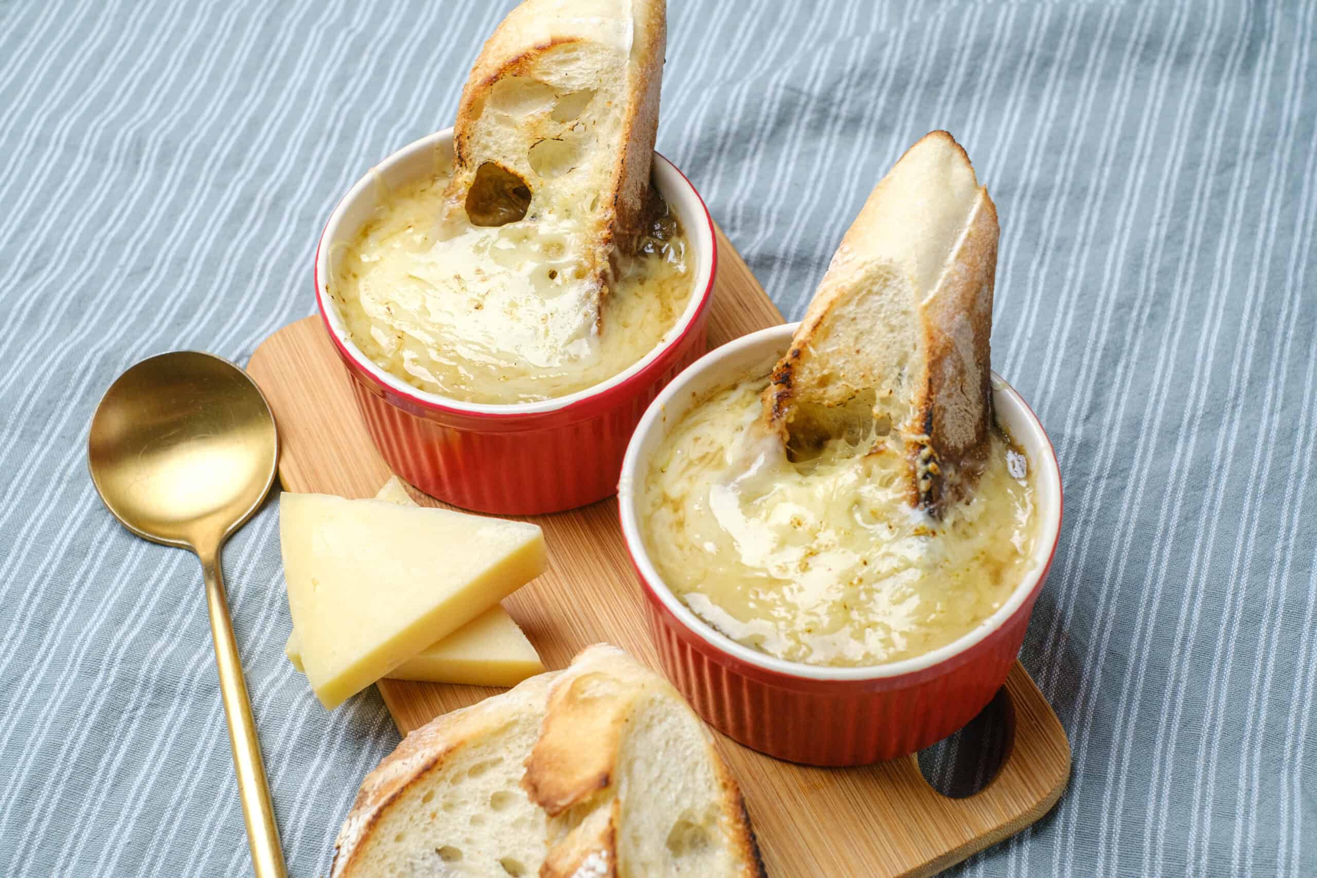 Creamy French onion soup served in red ramekins with toasted baguette slices and melted cheese, garnished with sliced cheese on a wooden board, perfect for cozy comfort food.