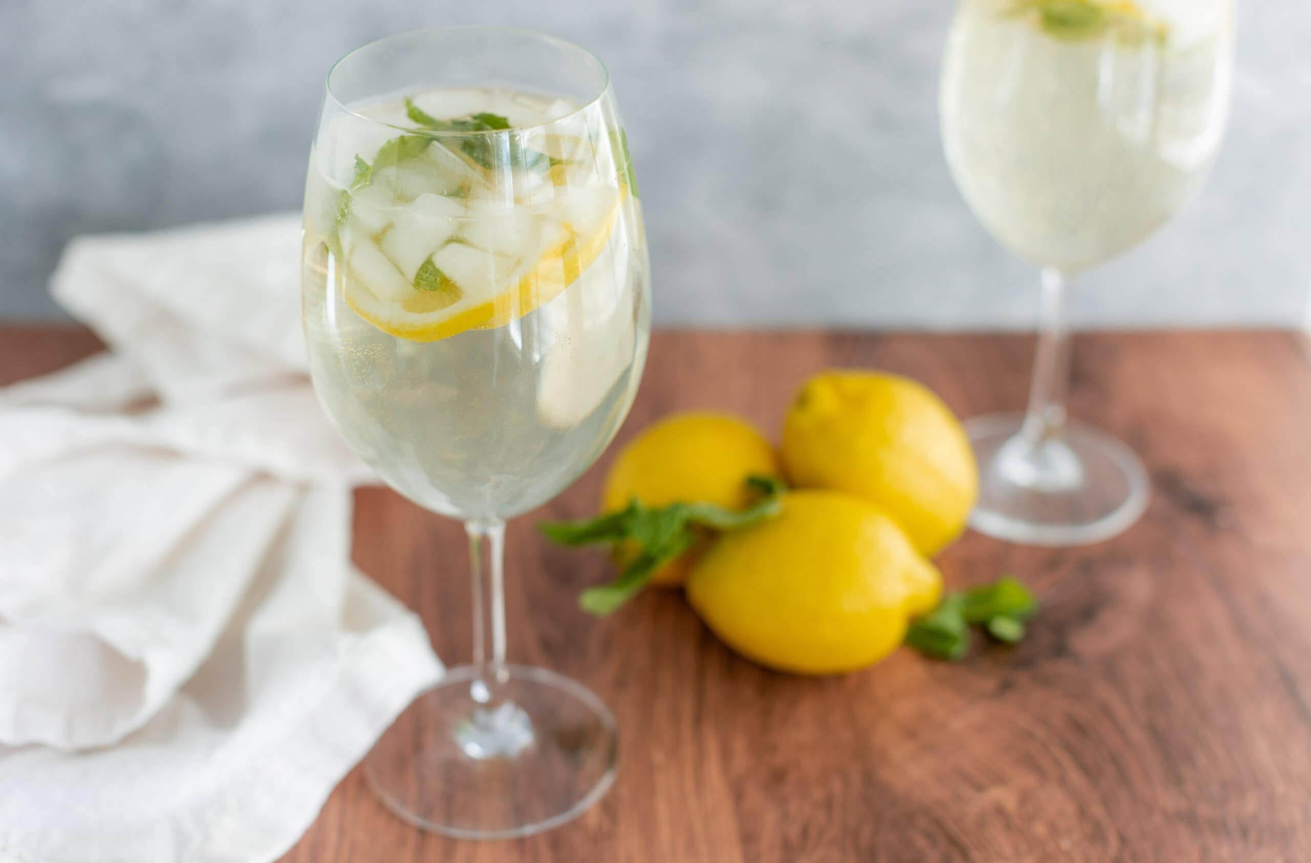 Refreshing lemon and cucumber sparkling water in elegant wine glasses, garnished with fresh lemon slices and mint leaves, served on a rustic wooden table with whole lemons nearby.