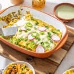 Colorful baked casserole with eggs, radishes, and green onions topped with melted cheese in a rustic orange baking dish on a wooden cutting board.