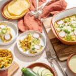 Savory breakfast casserole topped with cheese and sliced radishes, served with side dishes of corn, zucchini, and herb sauce on a rustic table with pink cloth napkins and vintage forks.