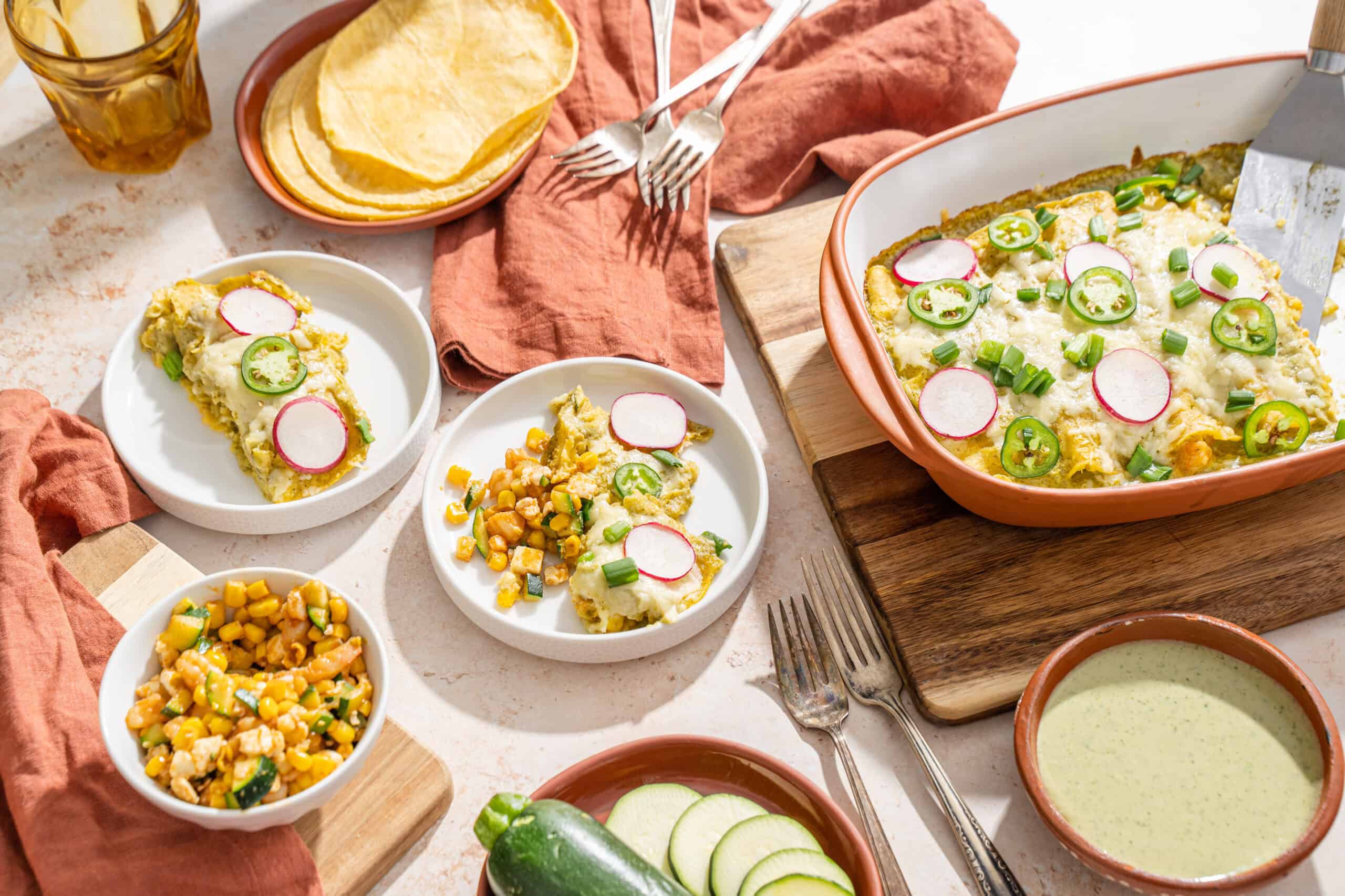 Savory breakfast casserole topped with cheese and sliced radishes, served with side dishes of corn, zucchini, and herb sauce on a rustic table with pink cloth napkins and vintage forks.