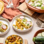 Creamy vegetable casserole topped with radish and green onion garnishes, served on white plates with fresh zucchini, corn, and tortilla chips - delicious vegetarian comfort food.
