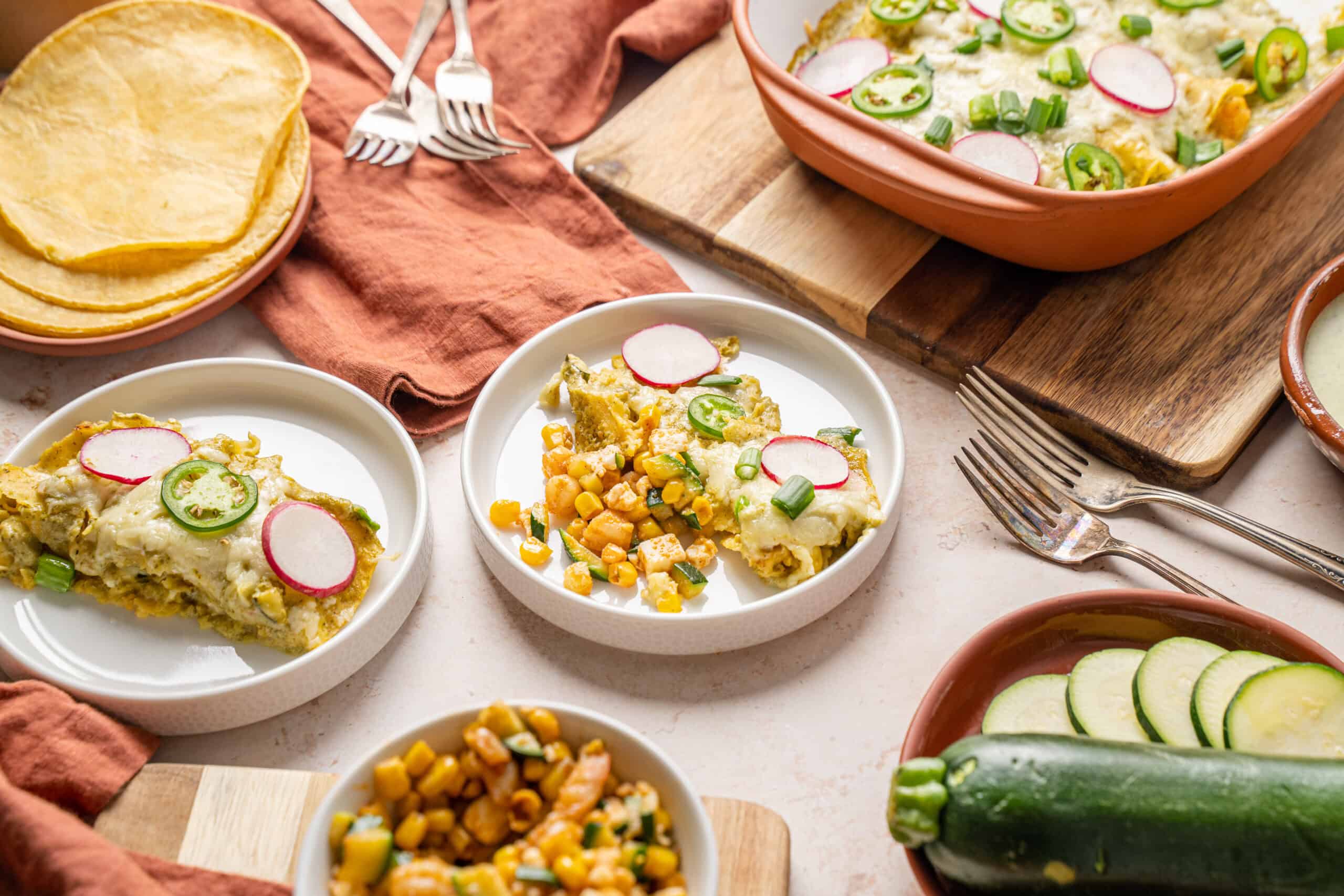Creamy vegetable casserole topped with radish and green onion garnishes, served on white plates with fresh zucchini, corn, and tortilla chips - delicious vegetarian comfort food.