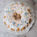 Colourful sprinkle-covered cake with white frosting on a glass plate, surrounded by scattered sprinkles and a metal knife, perfect for celebrations and dessert recipes.