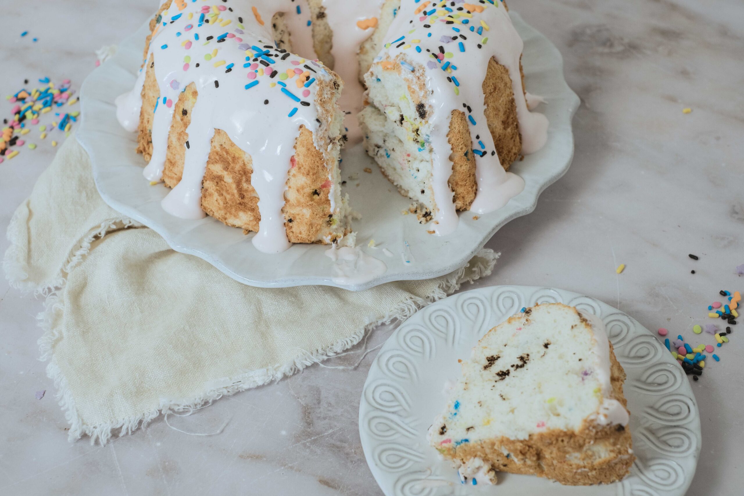 Vanilla cake with colorful sprinkles, topped with white icing and rainbow sprinkles, served on a decorative glass cake stand with a slice on a matching plate, on a marble surface with scattered sprinkles.