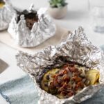 Aluminum foil-wrapped baked potato topped with crispy bacon, onions, and cheese, ready to serve; perfect for a hearty comfort food meal.