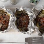 Baked stuffed potatoes with bacon, mushrooms, and cheese wrapped in foil on a white surface.