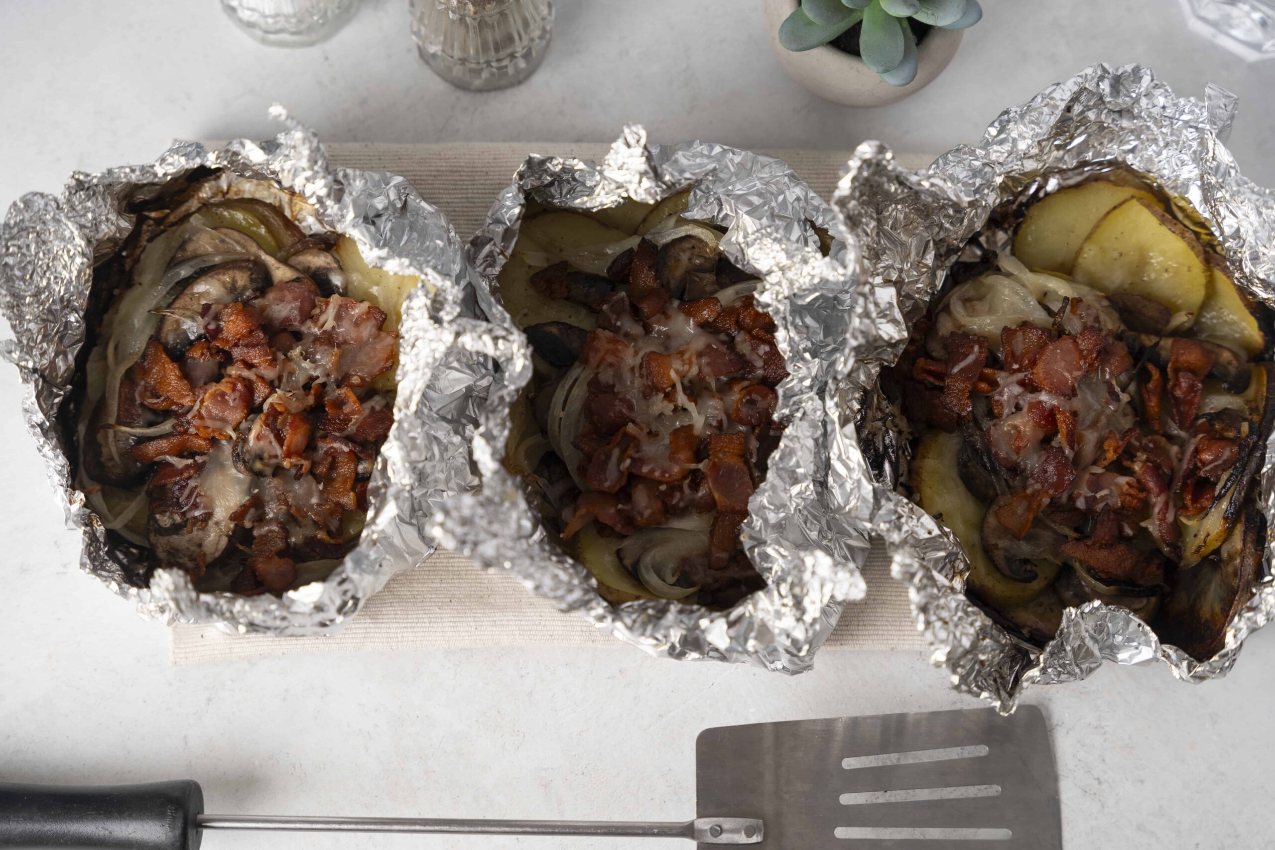 Baked stuffed potatoes with bacon, mushrooms, and cheese wrapped in foil on a white surface.
