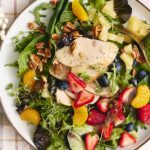 Fresh garden salad with grilled chicken, mixed greens, strawberries, blueberries, mandarin oranges, walnuts, and creamy vinaigrette dressing on a white plate.