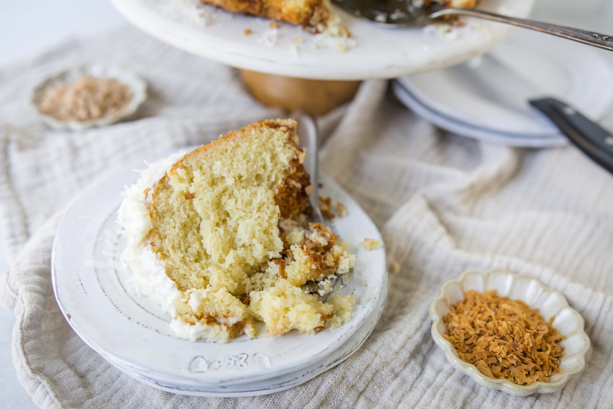 Rich, moist coconut cake with creamy frosting and toasted coconut flakes on top, served on a vintage white plate with a rustic cafe vibe, perfect for dessert or special occasions.
