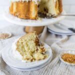 Fluffy homemade sponge cake with coconut frosting on a white cake stand, with a slice plated, garnished with toasted coconut flakes, on a rustic linen cloth.