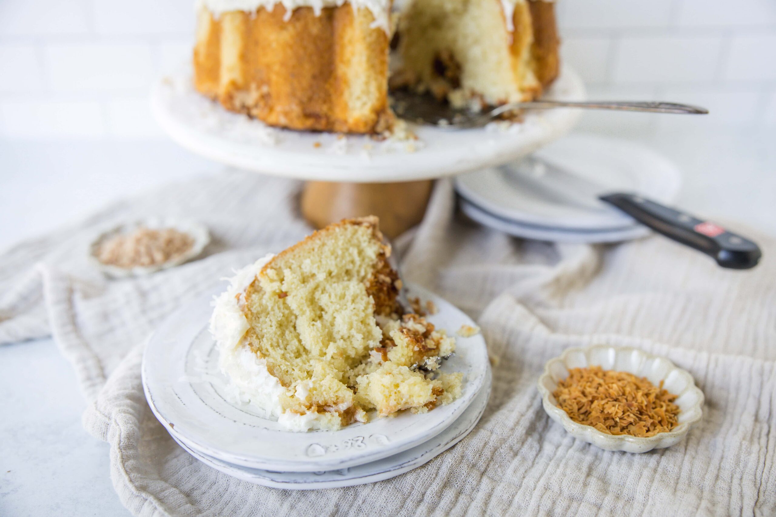 Fluffy homemade sponge cake with coconut frosting on a white cake stand, with a slice plated, garnished with toasted coconut flakes, on a rustic linen cloth.