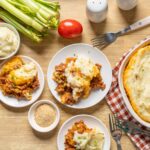 Baked shepherd's pie with mashed potatoes, ground beef, and vegetables on a wooden table with fresh veggies and serving dishes.