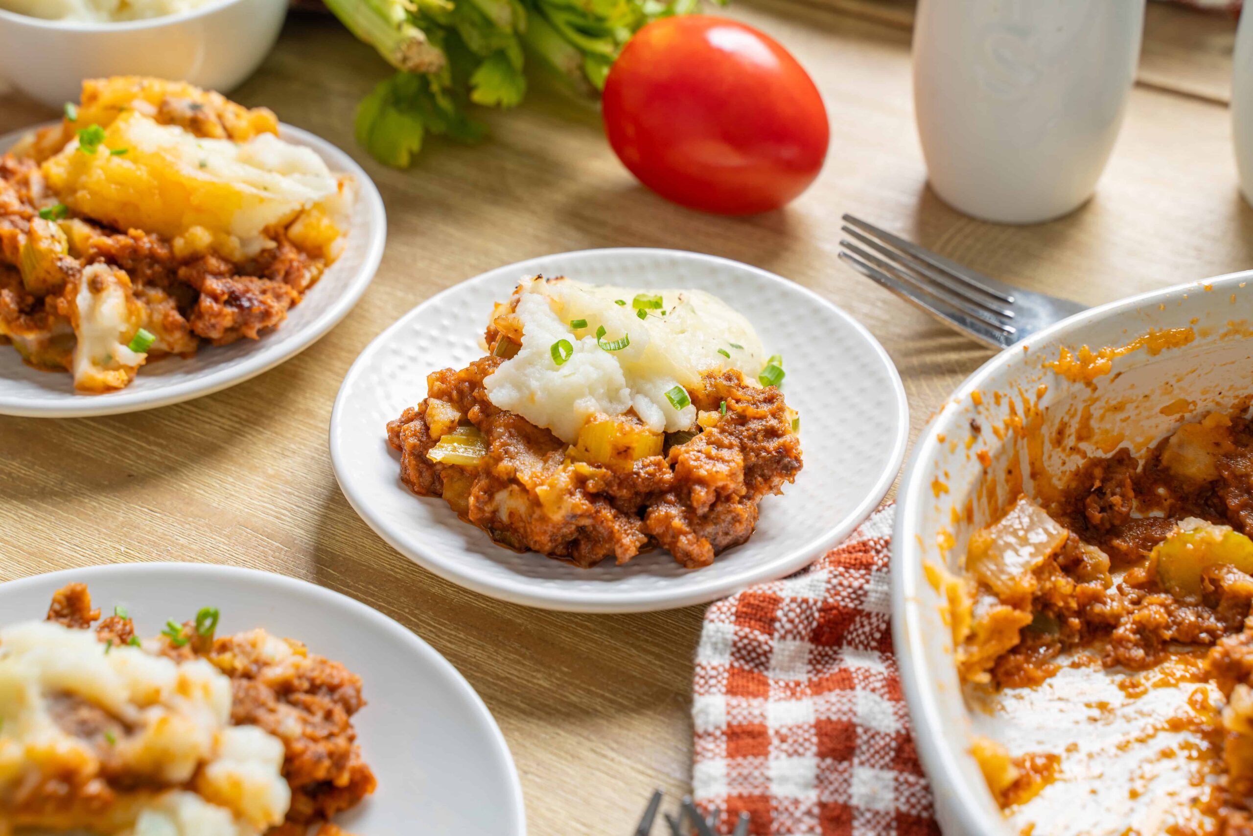 Savory homemade shepherd's pie with mashed potatoes and meaty beef filling served on white plates, fresh vegetables and utensils in the background; comforting winter dinner.