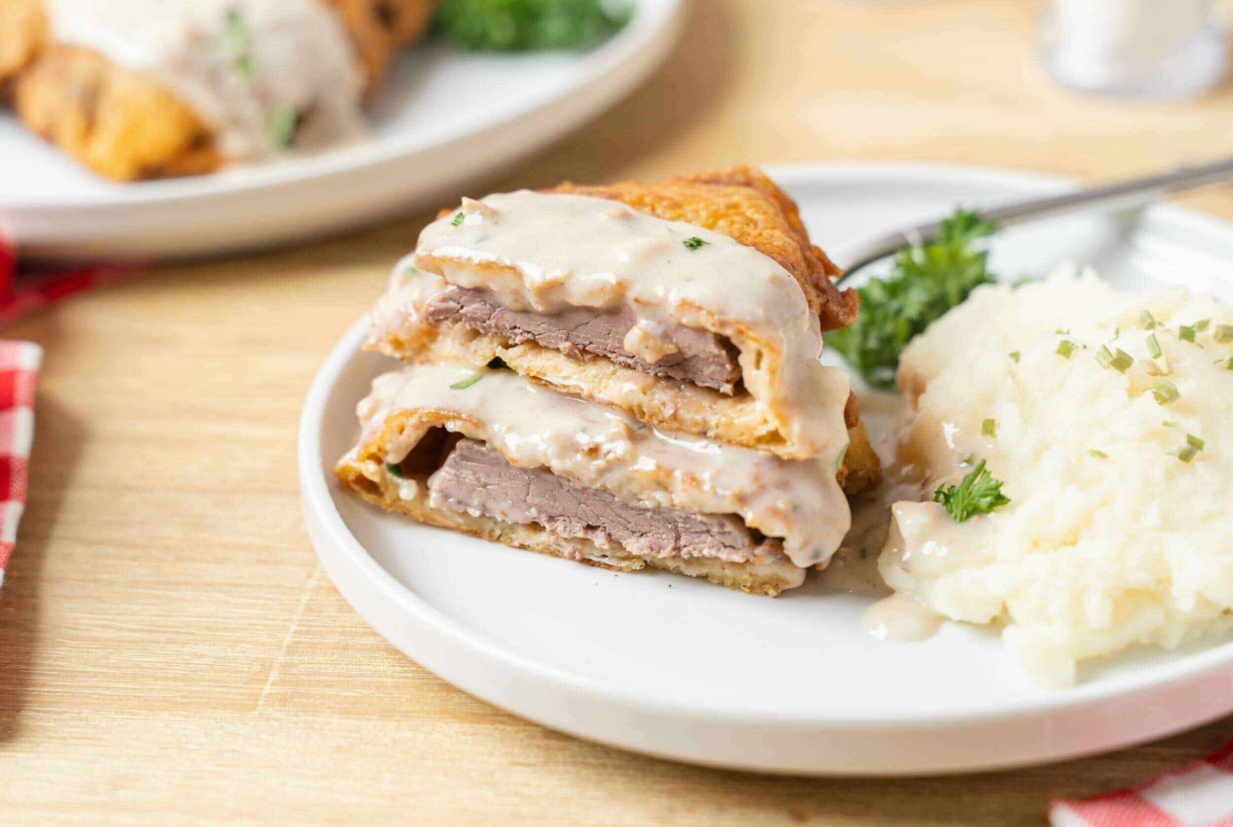 Thinly sliced beef steak and cheese rolling up in a golden, crispy egg batter with creamy white gravy. Served with mashed potatoes, parsley, and green herbs on a white plate.