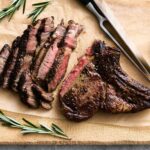 Juicy grilled steak with a perfectly seared crust and tender, pink interior on a wooden cutting board, garnished with fresh rosemary and served with black peppercorns.