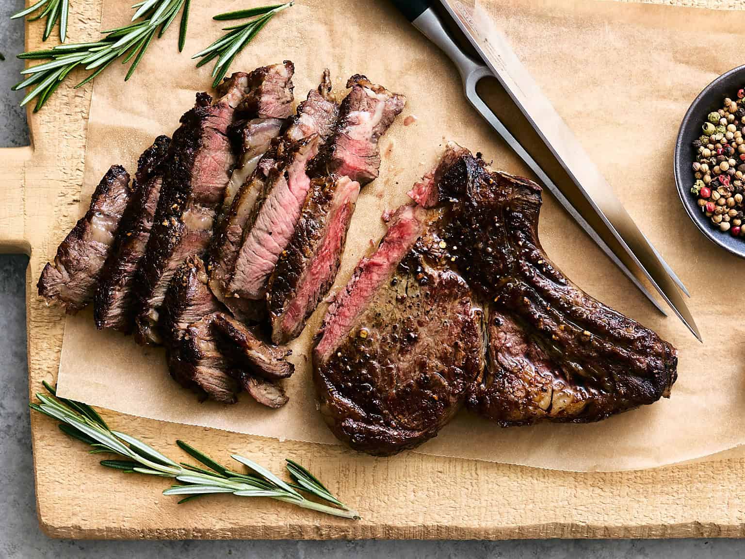 Juicy grilled steak with a perfectly seared crust and tender, pink interior on a wooden cutting board, garnished with fresh rosemary and served with black peppercorns.