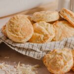 Buttery homemade cheese scones on a checkered cloth with shredded cheese on the side, perfect for breakfast or snack. Freshly baked, golden, and flaky scones made with cheese for a delicious treat.