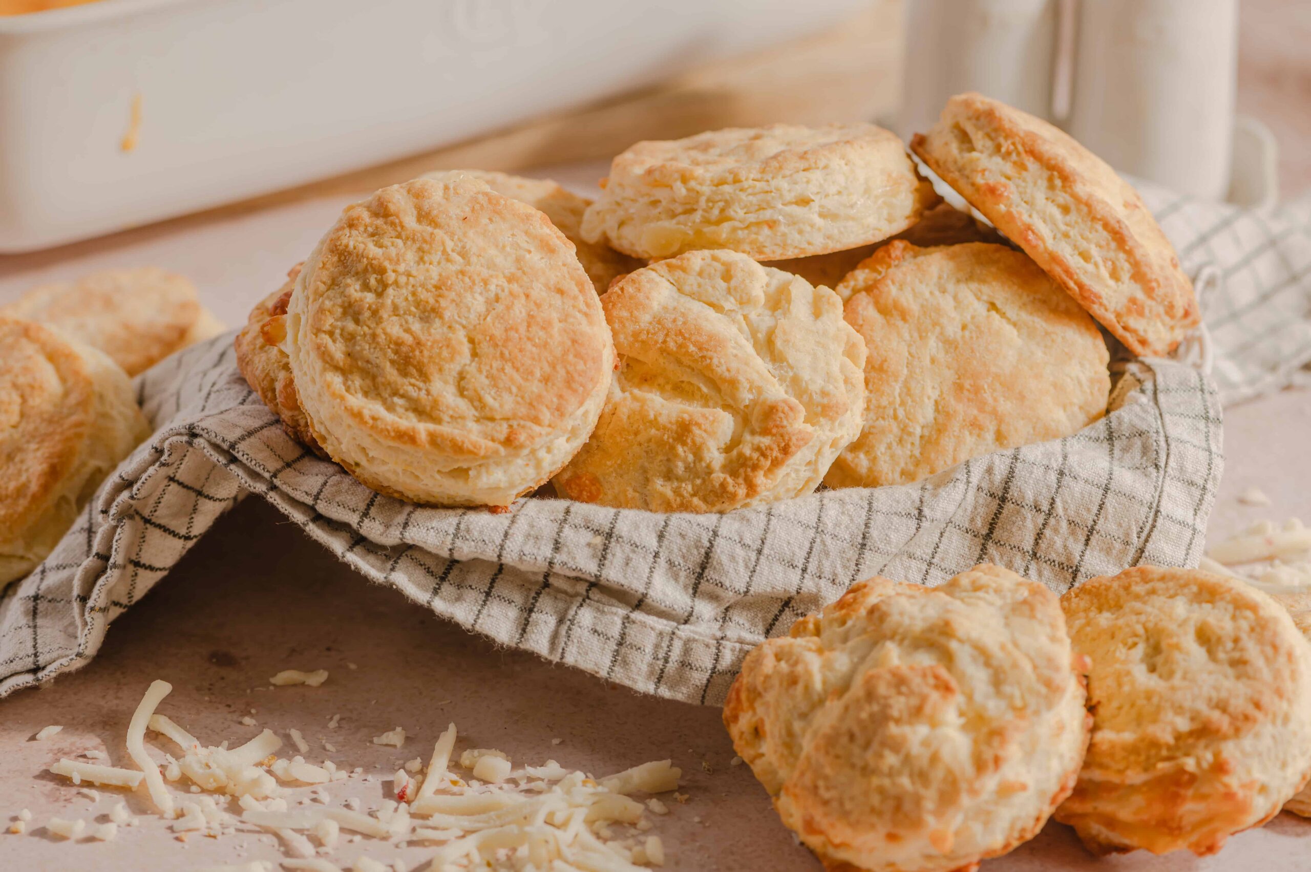 Buttery homemade cheese scones on a checkered cloth with shredded cheese on the side, perfect for breakfast or snack. Freshly baked, golden, and flaky scones made with cheese for a delicious treat.