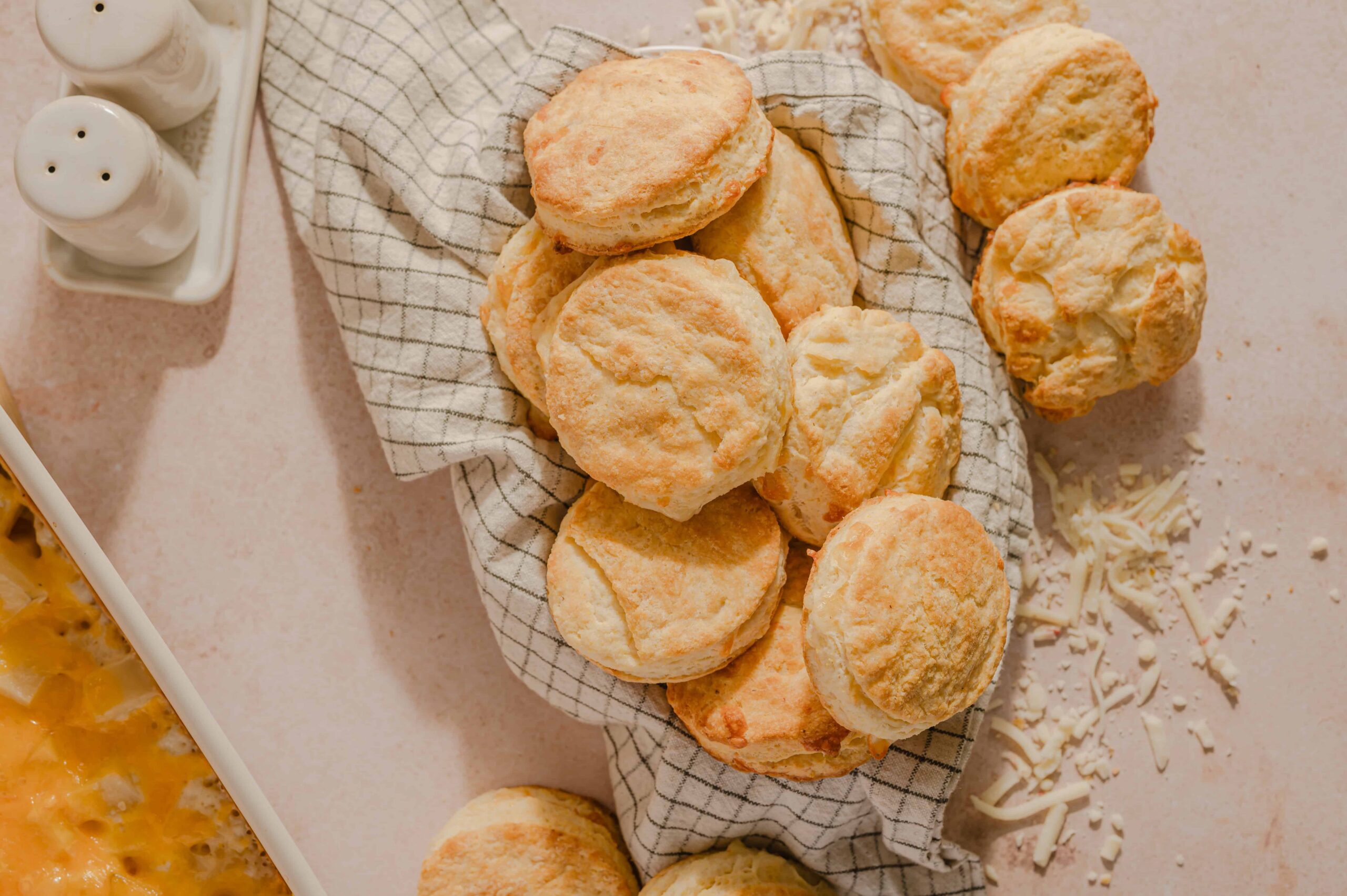 Fluffy homemade biscuits stacked on a checkered cloth, with grated cheese and seasoning nearby, ready to enjoy as a delicious snack or side dish. Perfect for breakfast or dinner, showcasing fresh baking.