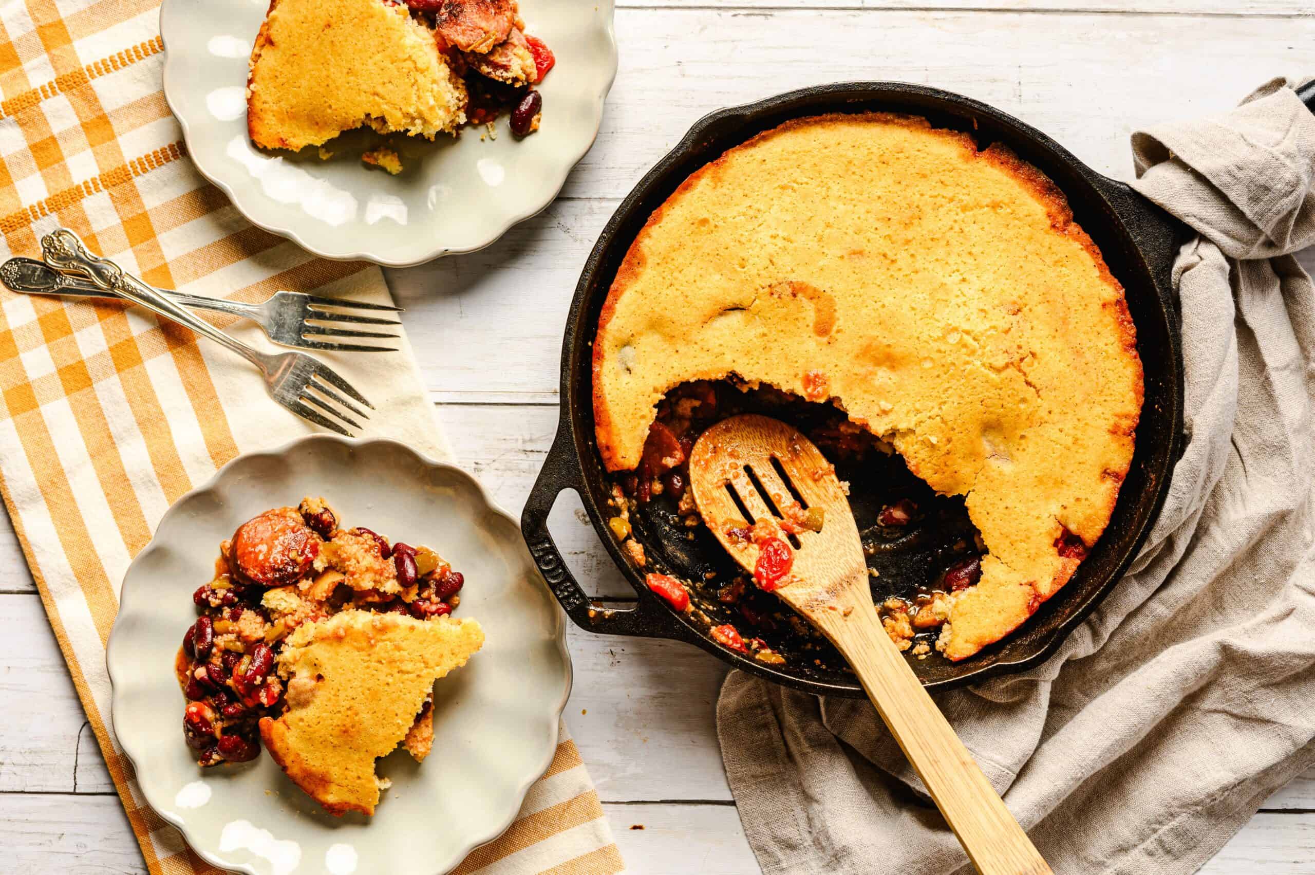 Rich, golden cornbread casserole with bean and sausage filling served in a cast iron skillet, with slices on decorative plates, perfect comfort food for family dinner.