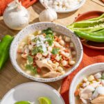 Fresh chicken and bean soup with chickpeas, topped with herbs, served in rustic bowls surrounded by fresh vegetables and limes for a flavorful Mexican-inspired dish.