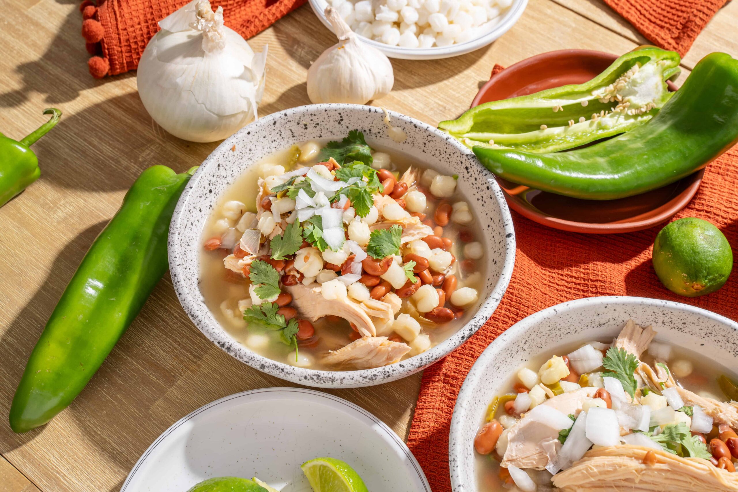 Fresh chicken and bean soup with chickpeas, topped with herbs, served in rustic bowls surrounded by fresh vegetables and limes for a flavorful Mexican-inspired dish.
