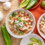 Fresh chicken tortilla soup garnished with beans, onions, and cilantro in rustic bowls, surrounded by garlic, lime, and colorful peppers on a wooden table.