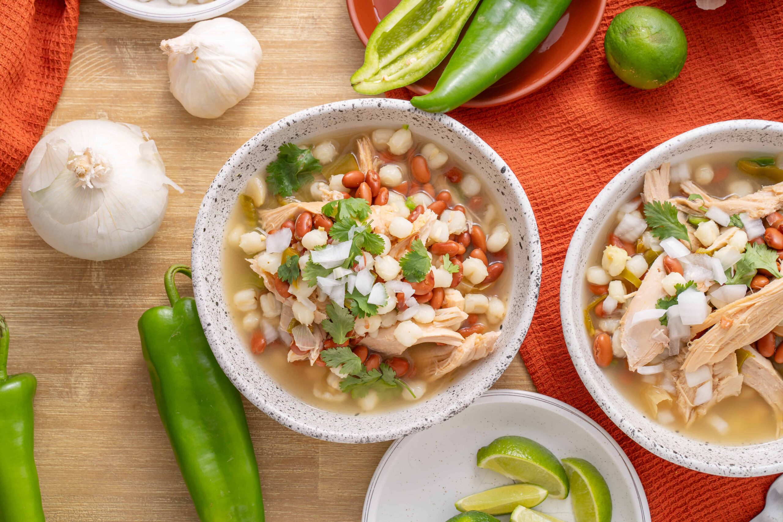Fresh chicken tortilla soup garnished with beans, onions, and cilantro in rustic bowls, surrounded by garlic, lime, and colorful peppers on a wooden table.