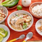 Succulent Mexican chicken soup with fresh herbs and beans in rustic bowls on an orange tablecloth, featuring lime wedges and colorful vegetables for homemade comfort food.
