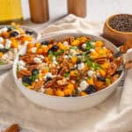 Savory holiday farro salad with roasted sweet potatoes, pecans, dried cranberries, feta cheese, and fresh herbs in a white serving bowl, styled for a cozy, rustic kitchen setting.