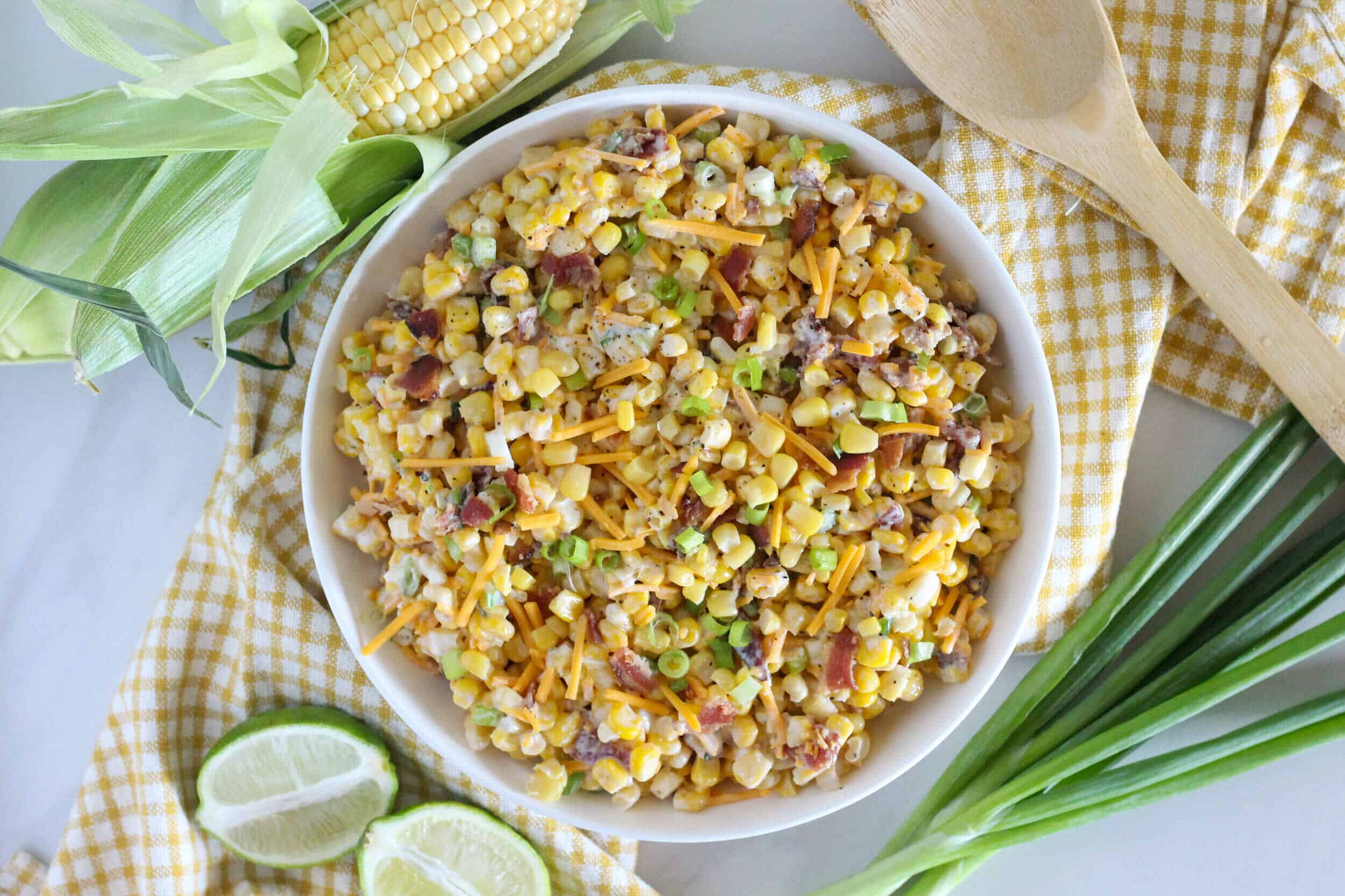 Corn chowder salad with bacon and green onions on a white bowl, served with lime slices, fresh corn, and green onions, perfect for summer barbecue or picnic meals, hearty and flavorful dish.