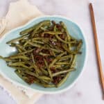Sauteed green beans with bacon on a light blue plate, garnished with cooked bacon bits, served on a cream-colored cloth, with wooden chopsticks on the side, ideal for a tasty vegetable side dish.