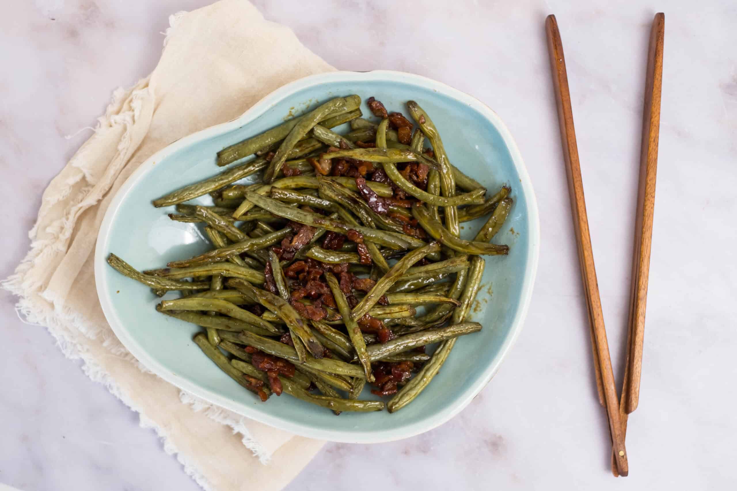 Sauteed green beans with bacon on a light blue plate, garnished with cooked bacon bits, served on a cream-colored cloth, with wooden chopsticks on the side, ideal for a tasty vegetable side dish.