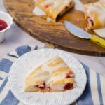 Fluffy cherry and cream cheese crepe cake slice served on a white decorative plate with a blue striped napkin, with a wooden serving board and a small bowl of cherry sauce in the background.