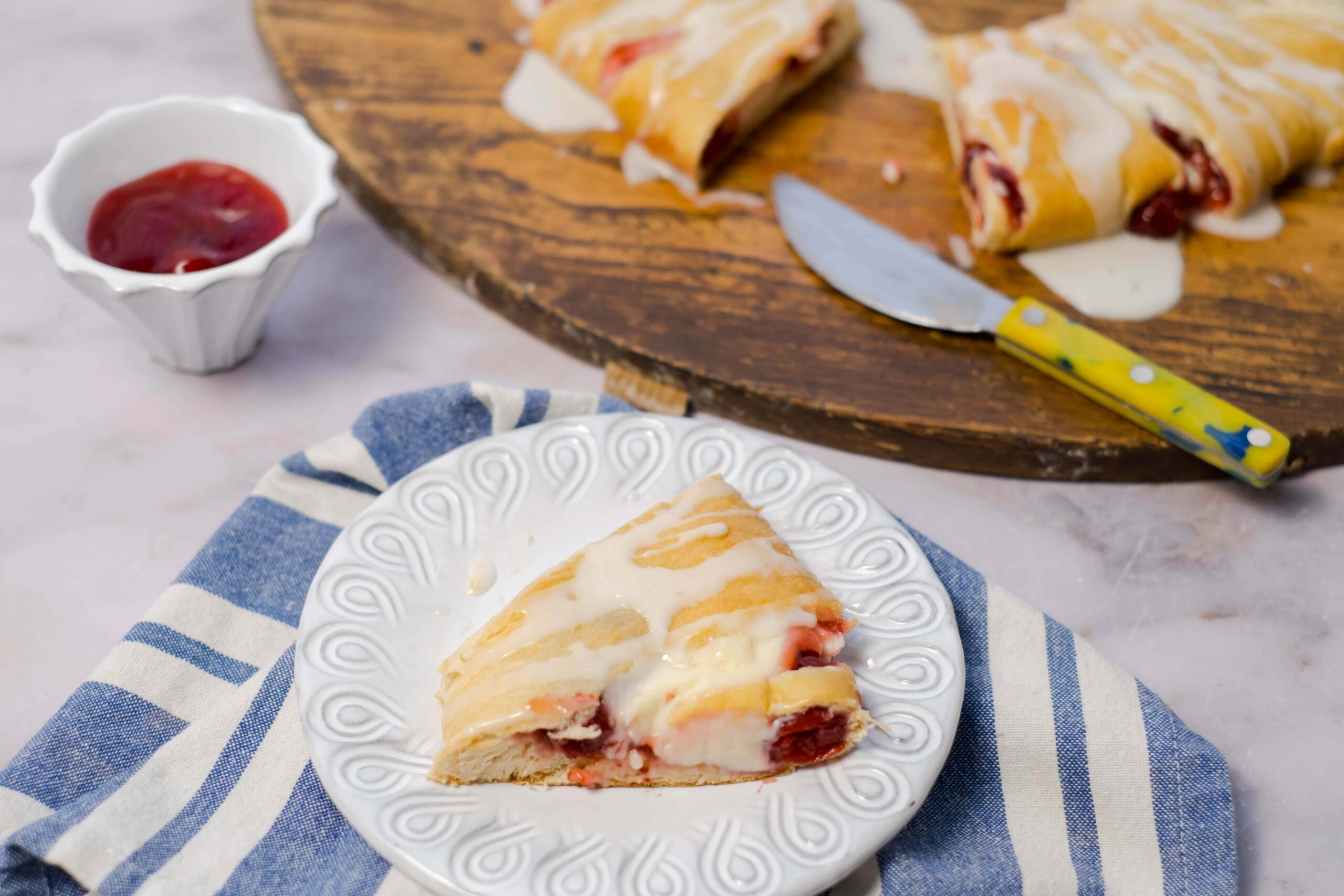 Fluffy cherry and cream cheese crepe cake slice served on a white decorative plate with a blue striped napkin, with a wooden serving board and a small bowl of cherry sauce in the background.