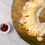 Freshly baked strawberry Danish pastry drizzled with icing on a rustic wooden board with a small bowl of strawberry jam and a blue striped towel on a white marble surface.