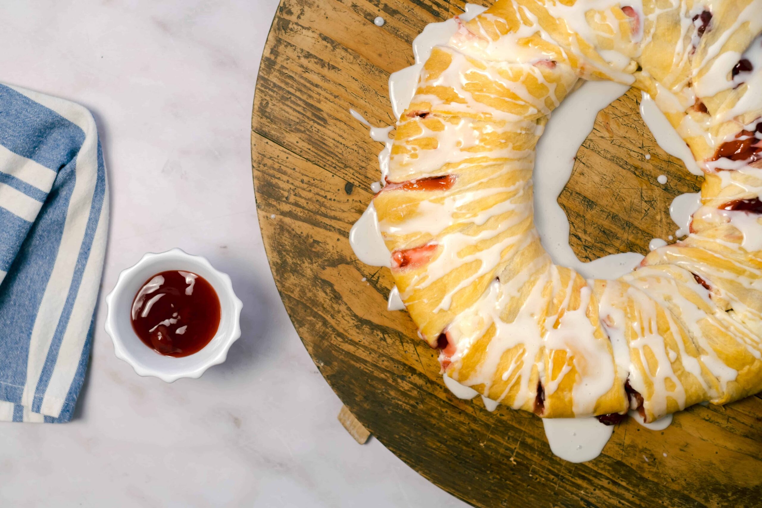Freshly baked strawberry Danish pastry drizzled with icing on a rustic wooden board with a small bowl of strawberry jam and a blue striped towel on a white marble surface.