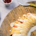 Golden brown croissant drizzled with white icing on a rustic wooden serving board, served with a side of strawberry jam and a lemon-yellow butter knife in the background.