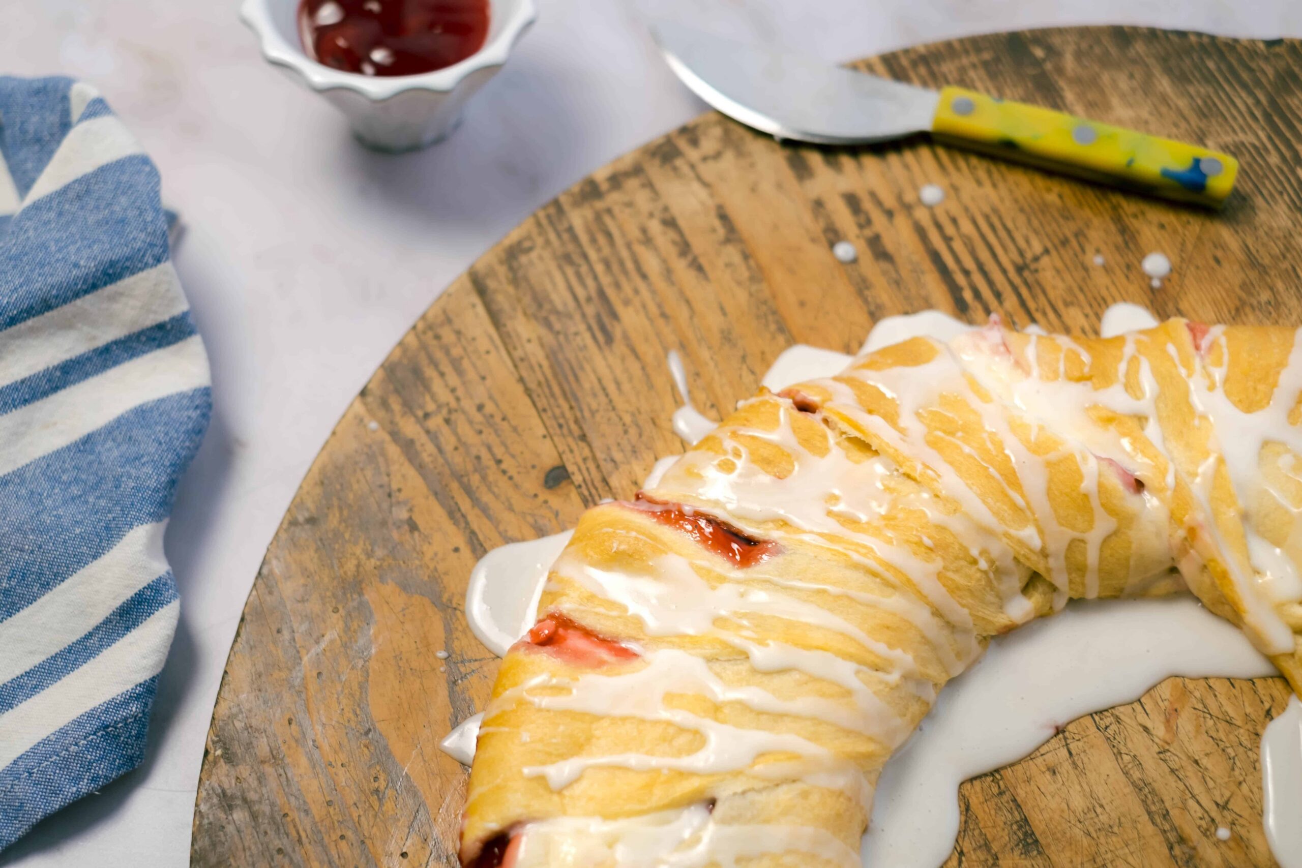 Golden brown croissant drizzled with white icing on a rustic wooden serving board, served with a side of strawberry jam and a lemon-yellow butter knife in the background.