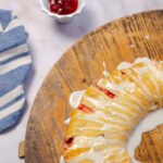 Sweet breakfast croissant drizzled with white icing on a rustic wooden board with a small bowl of cherry jam nearby.