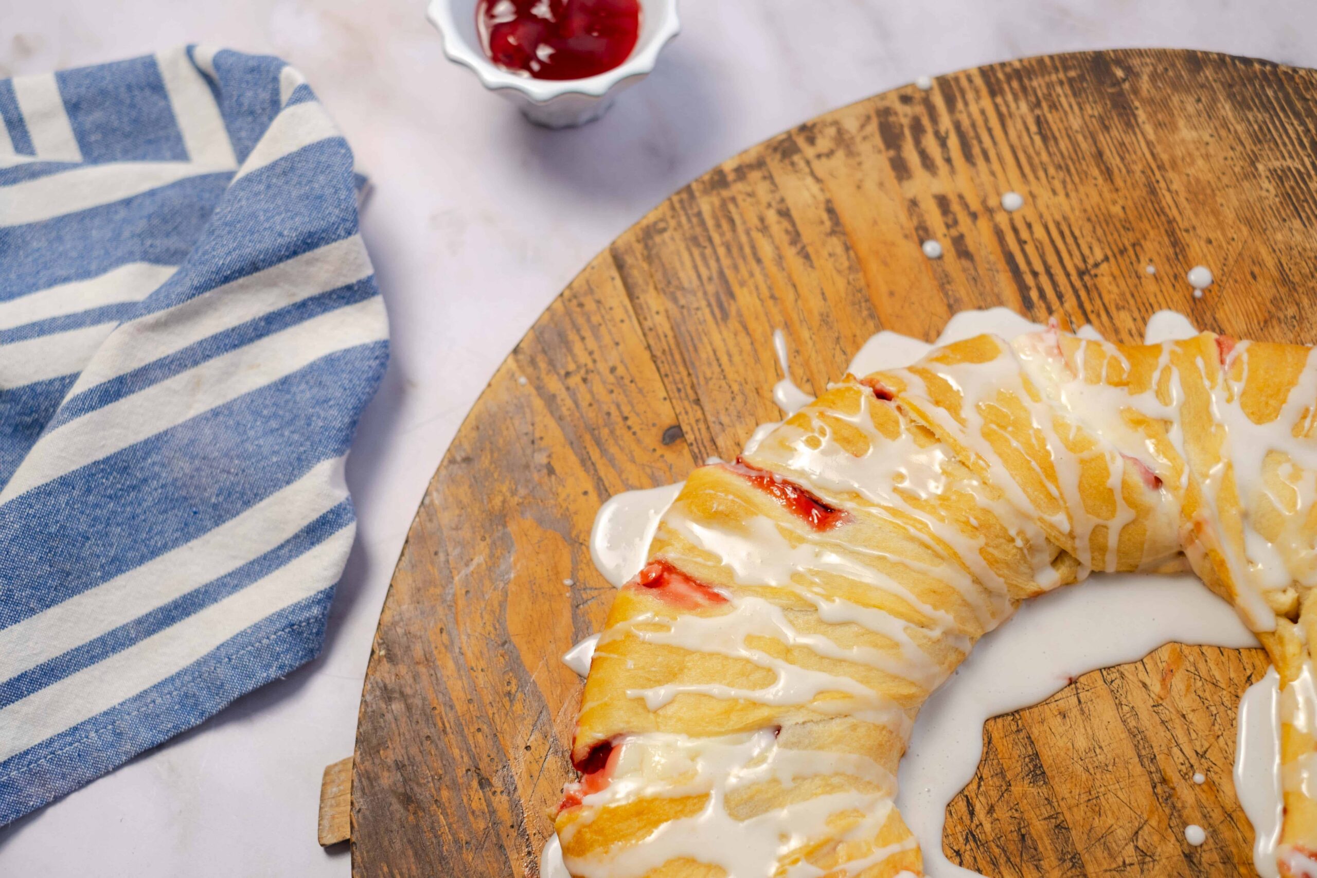 Sweet breakfast croissant drizzled with white icing on a rustic wooden board with a small bowl of cherry jam nearby.