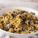 Creamy beef stroganoff with egg noodles, mushrooms, and herbs in a white bowl, served on a rustic table.