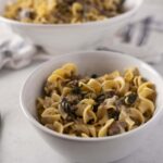 Creamy beef and mushroom tortellini pasta served in a white bowl with herbs, on a white table with a fork, water glass, and napkin for a delicious meal.