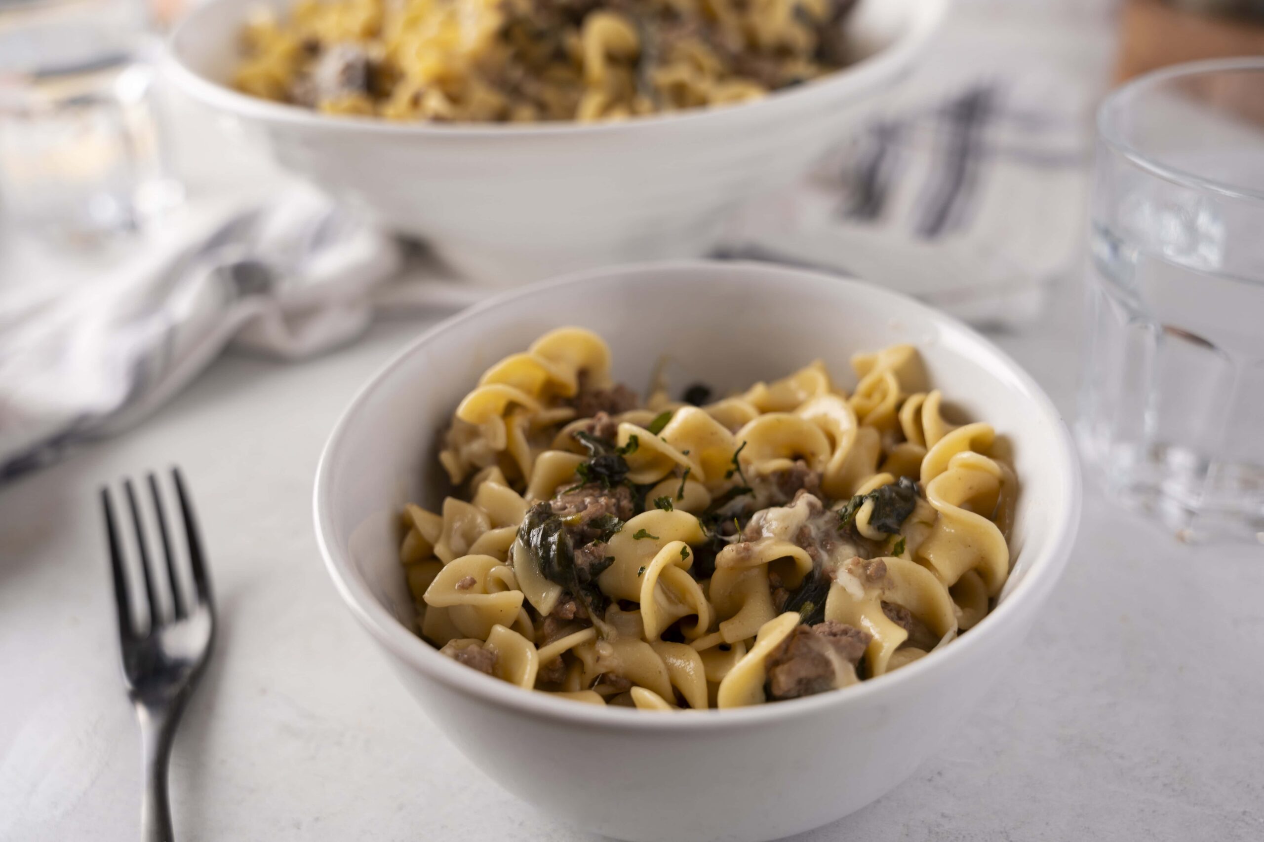 Creamy beef and mushroom tortellini pasta served in a white bowl with herbs, on a white table with a fork, water glass, and napkin for a delicious meal.