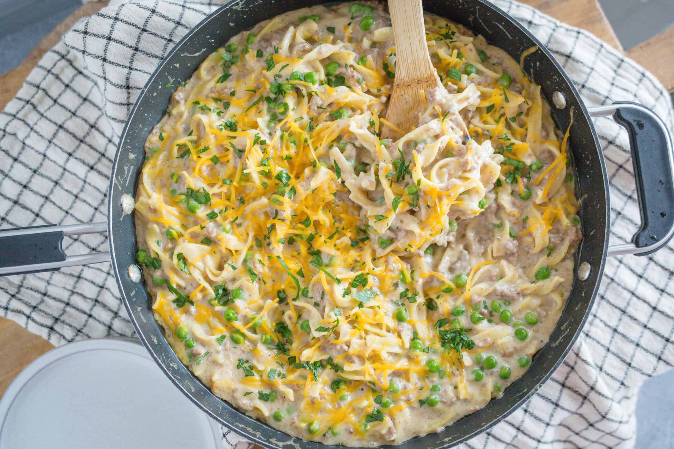 Creamy beef stroganoff with egg noodles, peas, and melted cheese garnished with parsley in a skillet. Perfect comfort food recipe for family dinners and easy weeknight meals.